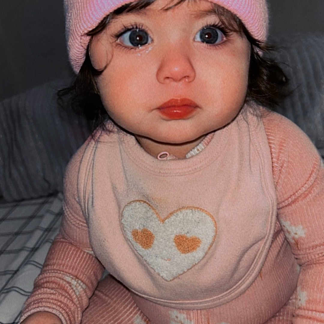Noah is registered to the contest to win money with this photo: baby, bed, bib, big_eyes, blanket, child, close_up, cozy_clothing, curly_hair, cute, heart_design, indoor, infant, long_sleeve, pants, pink_hat, pom_pom, portrait, sitting, warm_colors