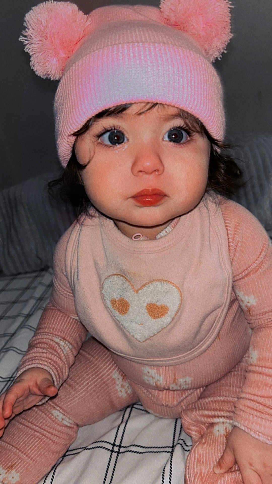 Noah is registered to the contest to win money with this photo: baby, child, pink_hat, pom_pom, cozy_clothing, long_sleeve, pants, bib, heart_design, indoor, bed, blanket, curly_hair, big_eyes, close_up, portrait, cute, sitting, infant, warm_colors