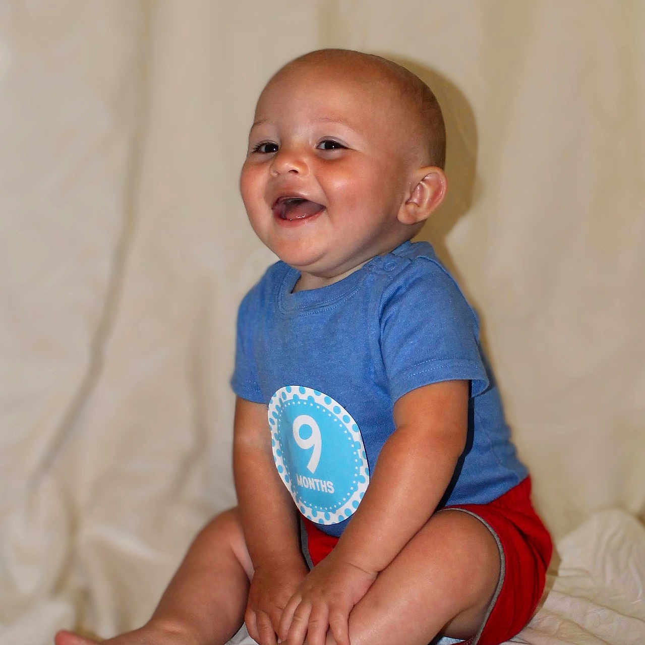 Kalub is registered to the contest to win money with this photo: baby, background, blue_shirt, child, clothing, cute, face, feet, hands, happy, indoor, infant, legs, nine_months, portrait, red_shorts, sitting, skin, smiling, young