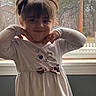 Ayah is registered to the contest to win money with this photo: child, girl, pigtails, white_dress, cat_print, bow, window, indoor, pose, playful, smile, face, hair, long_sleeves, expression, person, young_child, cute, casual_clothing, natural_light