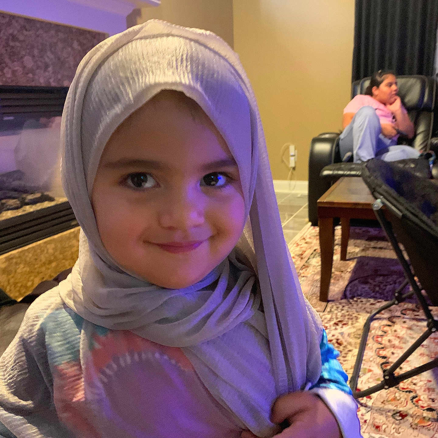 Ayah is registered to the contest to win money with this photo: child, hijab, smile, face, indoors, living_room, carpet, table, chair, person, adult, curtains, window, fireplace, rug, hand, clothing, casual, portrait, happy