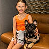 animal, brown, casual_clothing, child, companion, couch, dog, french_bulldog, friendly, girl, happy, indoor, leather_furniture, orange_tank_top, pet, portrait, shorts, sitting, smile, young