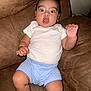 Julian is registered to the contest to win money with this photo: baby, child, infant, couch, brown_couch, white_shirt, blue_shorts, wide_eyes, surprised_expression, indoor, furniture, person, seated, hand_raised, cute, young_child, portrait, casual_clothing, skin, face