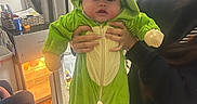 Sammy is registered to the contest to win money with this photo: baby, child, dinosaur_costume, green, person, indoor, carpet, mini_fridge, curtains, socks, hands, adult, clothing, face, cute, costume, holding, toy_story, tom_and_jerry, home