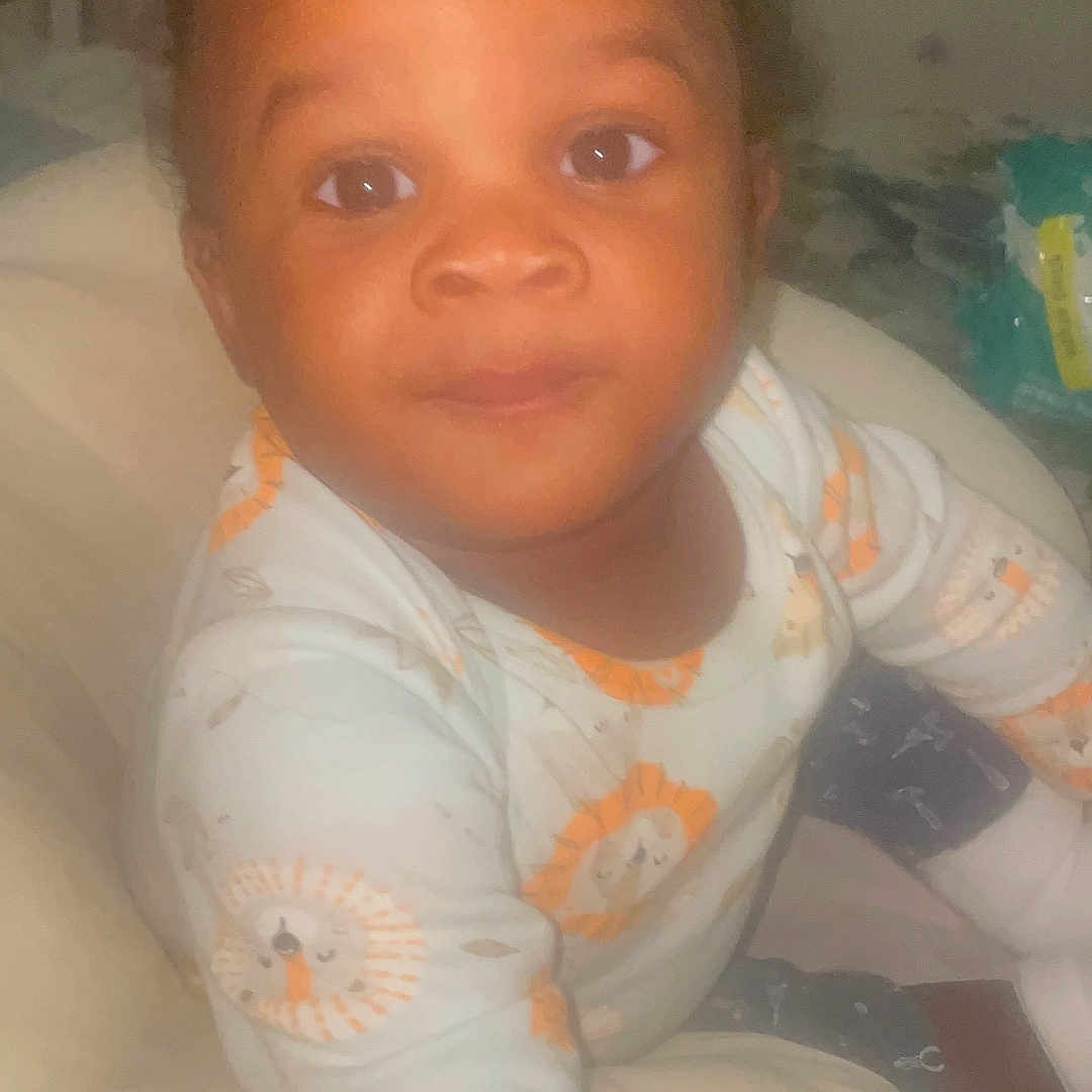 Nasir joined the competition — help win amazing prizes! baby, child, infant, onesie, lion_pattern, face, eyes, cushion, home_interior, furniture, soft_lighting, person, portrait, curious, cute, indoors, sleepwear, head, skin, smiling