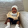 Edwin joined the competition — help win amazing prizes! baby, infant, smiling, sitting, couch, sofa, cushion, hat, beanie, socks, clothing, zip_jacket, t_shirt, text_on_clothing, dark_skin, happy, big_eyes, hands, indoor, window
