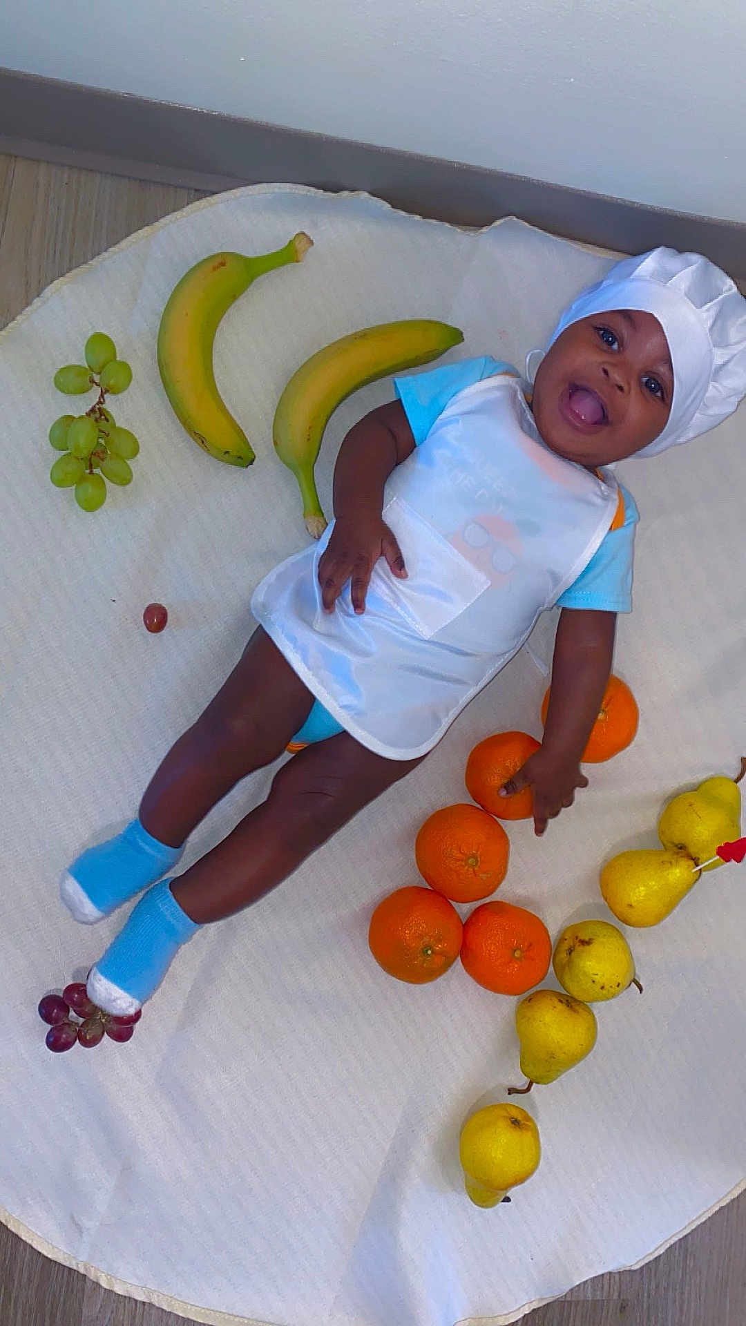 Edwin is registered to the contest to win money with this photo: child, baby, infant, smiling, chef_hat, bib, socks, bananas, grapes, oranges, pears, fruit, blanket, floor, lying_down, playful, happy, portrait, hand, feet