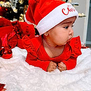 Naisha joined the competition — help win amazing prizes! baby, infant, christmas, santa_hat, red_clothing, blanket, holiday, decorations, christmas_tree, festive, cute, face, child, indoors, celebration, winter, smiling, holiday_lighting, soft_texture, toddler