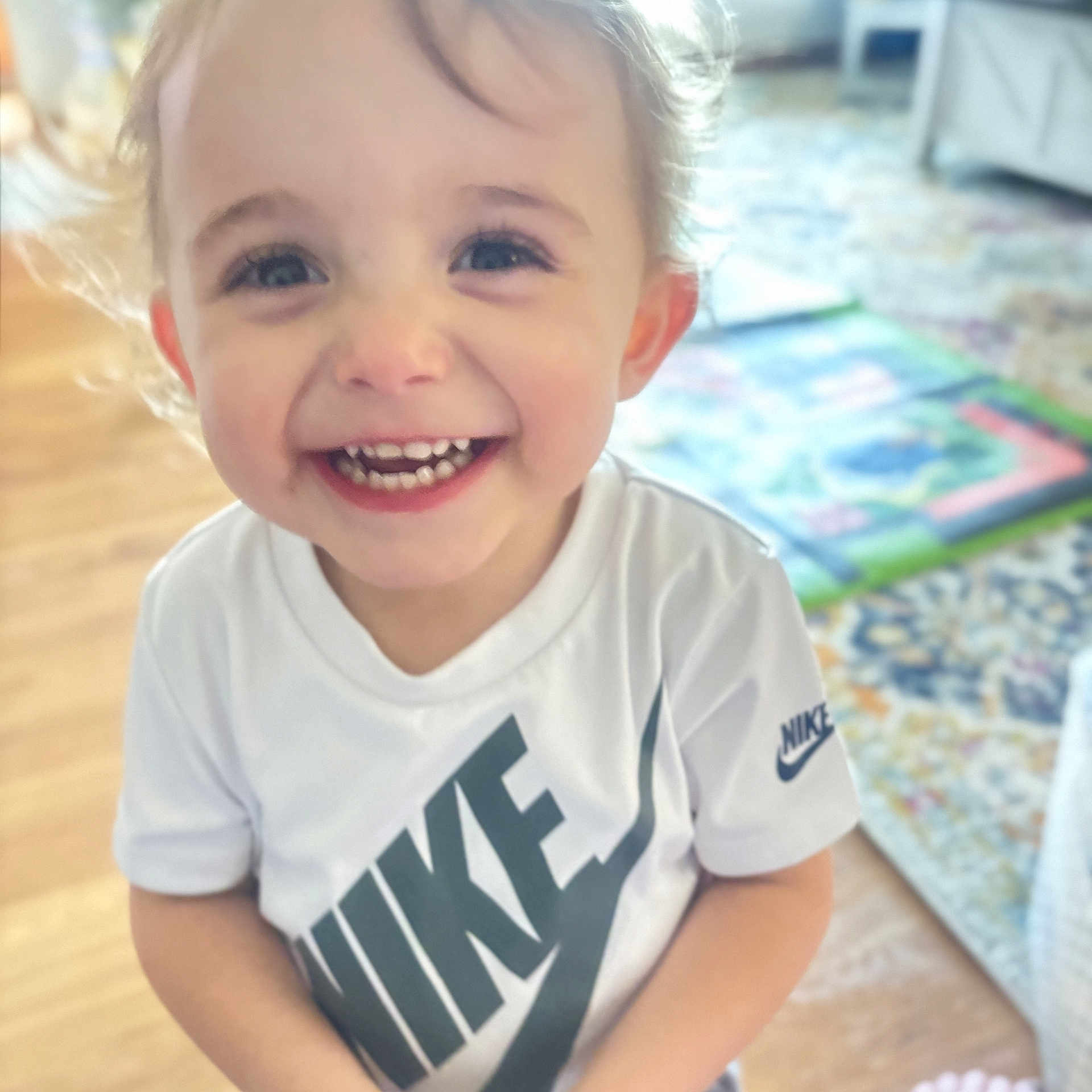 Alvin is registered to the contest to win money with this photo: child, toddler, smiling, happy, portrait, close_up, indoor, home, blond_hair, blue_eyes, teeth, white_shirt, nike_shirt, clothing, standing, play_mat, wood_floor, furniture, colorful_background, candid
