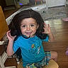 child, toddler, smiling, sitting, chair, long_hair, dark_hair, blue_shirt, plaid_shorts, indoor, living_room, wooden_floor, rug, toys, window, furniture, portrait, happy, playful, photography