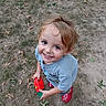 child, toddler, smiling, portrait, outdoor, grass, autumn, leaves, toy, red_toy, green_toy, playground, standing, gray_shirt, red_shoes, brown_hair, happy, looking_up, garden, daylight