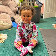 Brielle is registered to the contest to win money with this photo: toddler, child, smiling, floral_onesie, curly_hair, green_carpet, plush_teddy_bear, laundry_basket, clothes, toys, indoor, playroom, sitting, happy, cute, person, footed_pajamas, baby, carpet, home