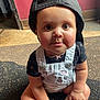 Brielle is registered to the contest to win money with this photo: baby, child, cap, denim_overalls, sitting, floor, indoor, curly_hair, wide_eyes, cute, light, shadow, red_wall, skin, hand, foot, bandage, black_shirt, expression, portrait