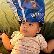 Priyah joined the competition — help win amazing prizes! baby, infant, child, face, portrait, sitting, lying_down, curious, cozy, cushion, green, clothing, pink_shirt, snack_bag, blue, funny_hat, indoor, home, close_up, humor