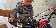 Tylio participe au concours pour gagner de l'argent avec cette photo : baby, child, motorbike, blue, garage, fishing_rod, concrete_wall, socks, adult_hand, tattoo, curious, grip, handlebar, footwear, floor, vehicle, helmet_absent, indoor, person, sitting