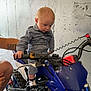 baby, child, motorbike, blue, garage, fishing_rod, concrete_wall, socks, adult_hand, tattoo, curious, grip, handlebar, footwear, floor, vehicle, helmet_absent, indoor, person, sitting