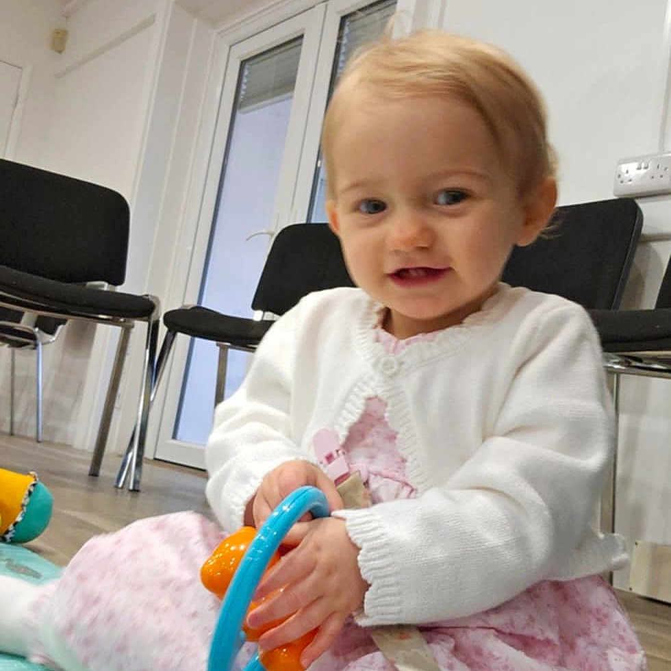 Alisha joined the competition — help win amazing prizes! baby, child, curly_hair, door, floor, furniture, happy, home, indoor, person, pink_dress, play_mat, playing, sitting, smiling, sock, toddler, toy, white_cardigan, window