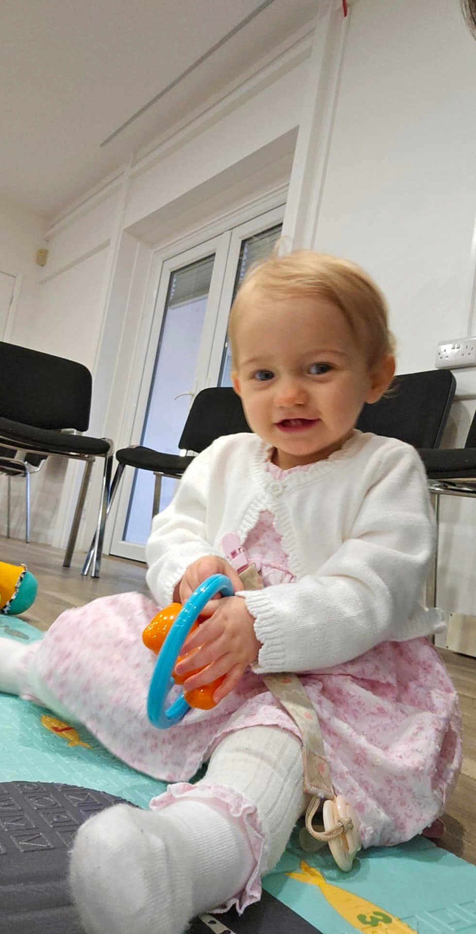 Alisha joined the competition — help win amazing prizes! toddler, child, indoor, play_mat, toy, white_cardigan, pink_dress, sitting, floor, smiling, curly_hair, sock, furniture, window, door, happy, person, baby, playing, home