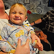 Alisha is registered to the contest to win money with this photo: toddler, child, smiling, floral_onesie, person, adult_hand, bracelets, living_room, couch, wallpaper, decor, happy, portrait, indoor, family, casual, seated, cozy, home, expression