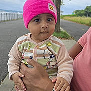 Sarah is registered to the contest to win money with this photo: adult_hand, baby, casual, child, clothing, curious_expression, cute, grass, holding, nature, outdoor, overcast_sky, person, pink_beanie, portrait, road, seated, sidewalk, tree, watch