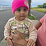 adult_hand, baby, casual, child, clothing, curious_expression, cute, grass, holding, nature, outdoor, overcast_sky, person, pink_beanie, portrait, road, seated, sidewalk, tree, watch
