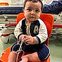 baby, child, orange_chair, medical_equipment, hospital_room, wire, bandage, foot, face, curly_hair, sitting, clothing, jacket, medical_monitor, floor, expression, indoors, person, cute, healthcare
