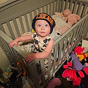 Sara-Ann is registered to the contest to win money with this photo: toddler, child, crib, beanie, hat, stuffed_animals, toys, blanket, clothing, indoor, wooden_floor, baby, person, furniture, cute, curious, gripping, standing, face, wide_eyes