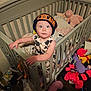 toddler, child, crib, beanie, hat, stuffed_animals, toys, blanket, clothing, indoor, wooden_floor, baby, person, furniture, cute, curious, gripping, standing, face, wide_eyes
