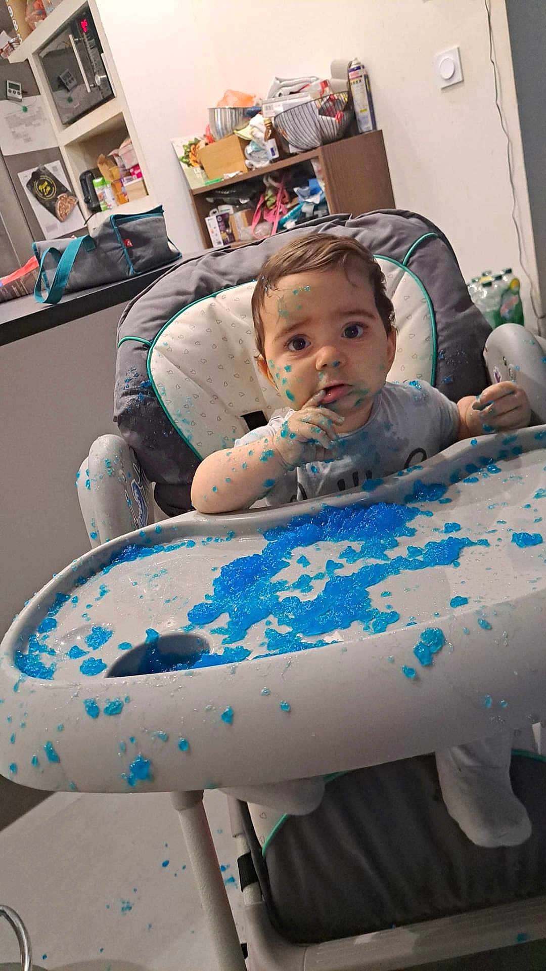 Léo participe au concours pour gagner de l'argent avec cette photo : baby, high_chair, blue_jelly, messy, food, kitchen, child, face, hands, tray, expression, indoor, curious, sitting, clothing, hair, footwear, table, background, household_items