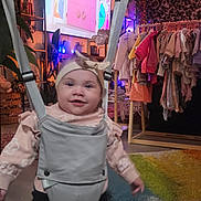 Roselie is registered to the contest to win money with this photo: baby, baby_toys, black_pants, child, clothes, clothing, colorful, floor, footwear, happy, headband, home, indoor, jumper, person, pink, plant, rack, smile, wood_floor