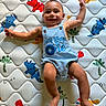 baby, infant, smiling, blue_romper, dinosaur_pattern, mattress, child, happy, cute, lying_down, arms_outstretched, colorful, playful, indoors, person, portrait, young_child, text_on_clothing, celebration, comfort