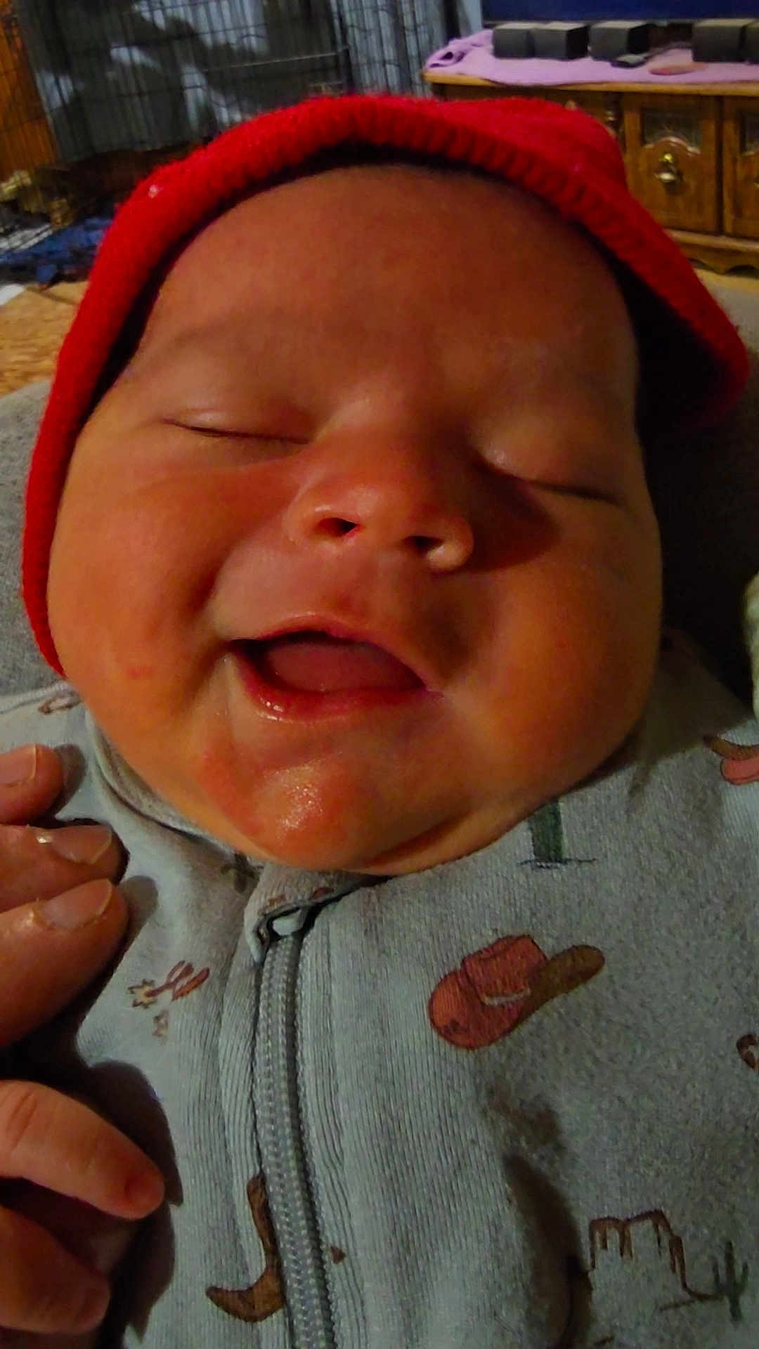 Oliver is registered to the contest to win money with this photo: baby, infant, smile, red_cap, close_up, hand, onesie, clothing, face, cheeks, mouth_open, sleepy_eyes, indoor, soft_lighting, newborn, cute, child, person, happy, portrait