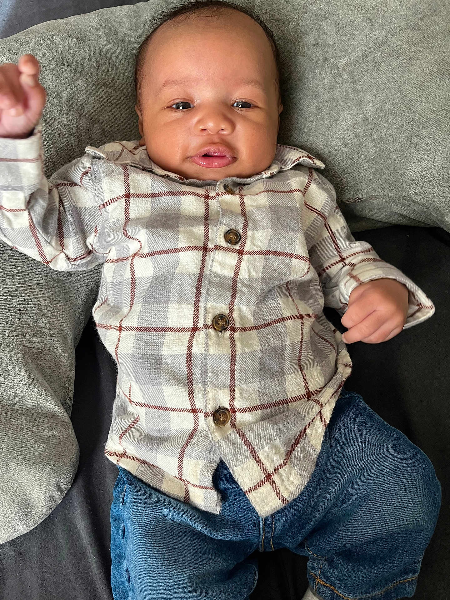 Legend is registered to the contest to win money with this photo: baby, child, plaid_shirt, blue_jeans, cushion, gray_cushion, infant, casual_clothing, indoor, person, cute, smiling, hand_raised, relaxed, portrait, young_child, comfort, happy, face, sitting