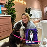 Coco is registered to the contest to win money with this photo: woman, dog, painting, indoor, pink_wall, plant, neon_sign, smile, carpet, chair, table, leash, portrait, artwork, pet, happy, blonde_hair, casual_clothing, decor, cozy