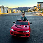 Luis joined the competition — help win amazing prizes! toddler, toy_car, red_car, child, street, sunset, suburban, houses, mountain, outdoor, asphalt, sky, residential_area, vehicle, head, face, driving, evening, side_mirror, road