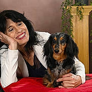 Sirena a rejoint le concours — aidez-le/la à gagner de superbes lots ! brown_background, casual, companion, dachshund, decorative_column, dog, friendly, greenery, happy, indoor, long_hair, pet, portrait, posing, red_tablecloth, resting_hand, smiling, studio_lighting, white_jacket, woman