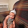 baby, child, infant, plaid_couch, striped_clothing, white_hat, toy, indoor, air_conditioner, sitting, curious, cozy, person, couch, home, cute, knit_hat, soft_lighting, young_child, focused