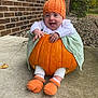 Miami is registered to the contest to win money with this photo: baby, pumpkin, knitted_hat, knitted_booties, orange, green_cloth, brick_wall, outdoor, sidewalk, rocky_garden, happy, smiling, child, cute, fall, seasonal, costume, sitting, person, portrait