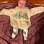 baby, infant, sweater, knitwear, blanket, lying_down, cute, newborn, clothing, person, child, soft_texture, warm_colors, maroon, orange, patterned_pants, face, head, expression, indoors
