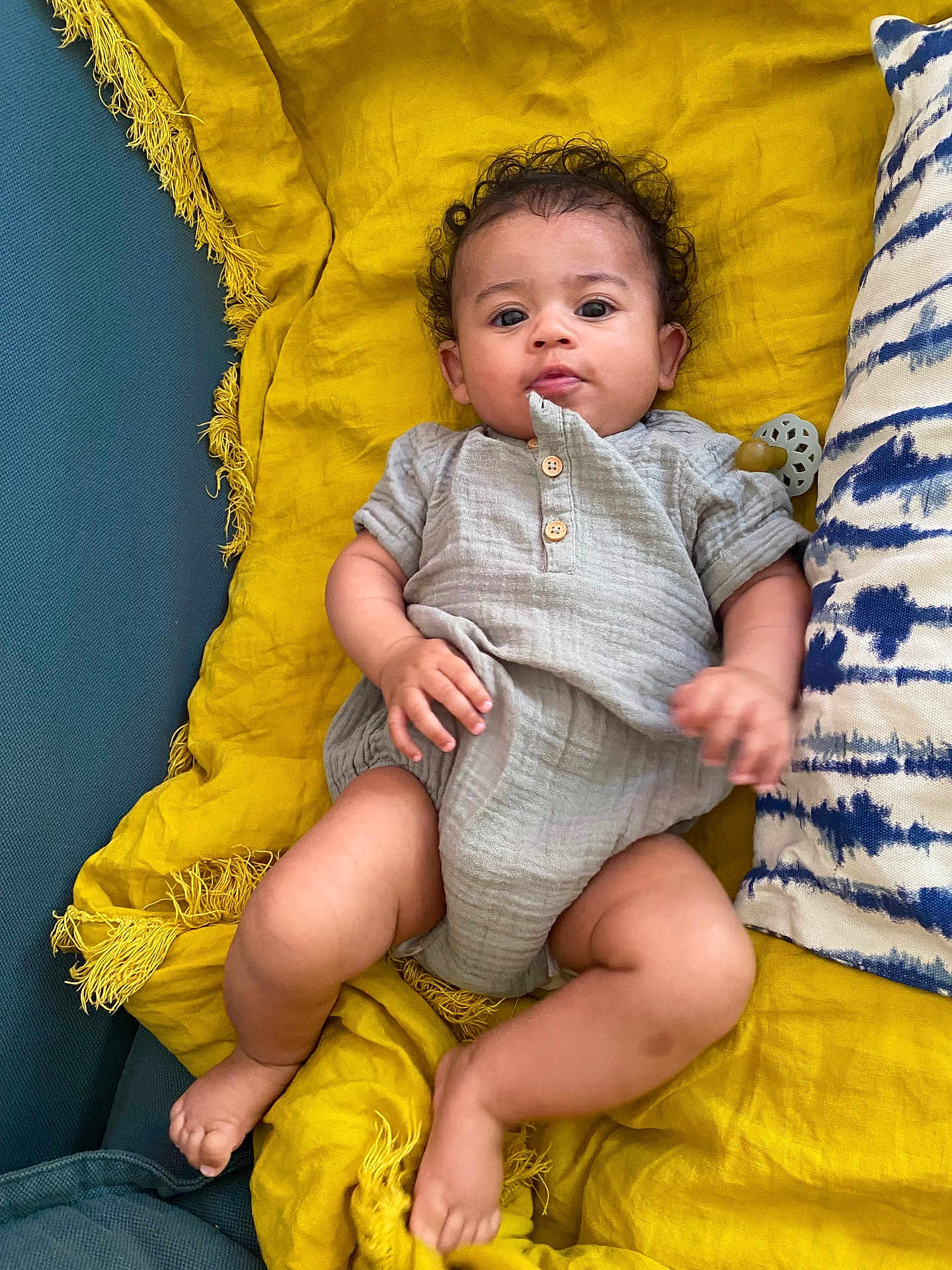 Mivia a rejoint le concours — aidez-le/la à gagner de superbes lots ! baby, infant, child, curly_hair, gray_outfit, yellow_blanket, blue_pillow, cushion, indoor, relaxed, lying_down, cute, soft_fabric, skin, hand, foot, expression, cozy, home, portrait