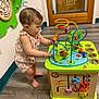 toddler, child, toy, bead_maze, indoor, wooden_floor, pacifier, barefoot, romper, plaything, colorful, curious, standing, wooden_door, wall_toy, flooring, person, baby, early_childhood, engagement