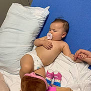 Arya is registered to the contest to win money with this photo: baby, pacifier, diaper, plush_doll, bed, pillow, hand, adult, toy, blue_bed_sheet, white_sheet, infant, sleeping, holding, skin, room, cable, comfort, child, lying_down