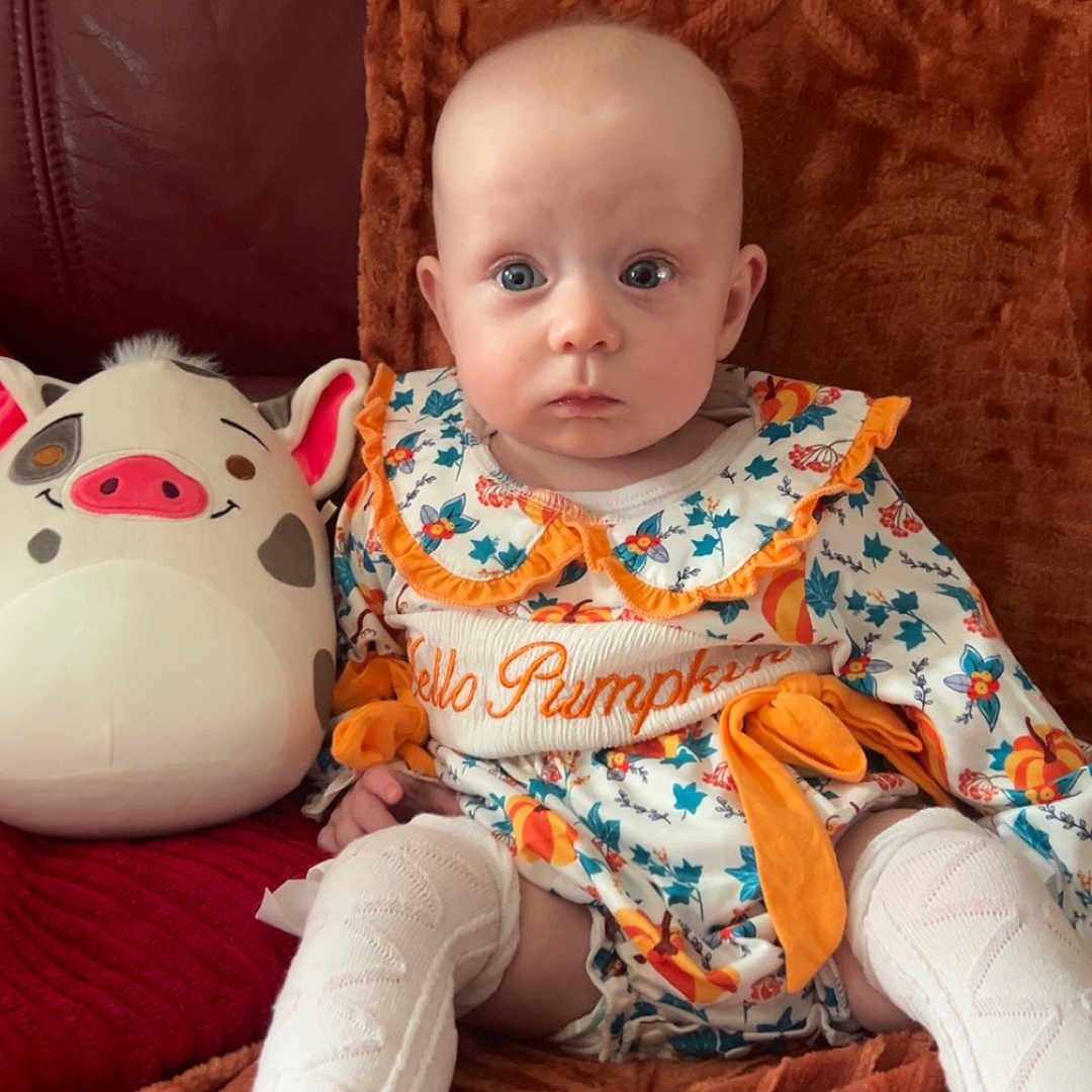 Eilidh joined the competition — help win amazing prizes! baby, blanket, child, colorful_clothing, couch, cozy, cute, expression, face, floral_pattern, indoor, infant, orange, plush_toy, portrait, seated, socks, soft_toy, white, wide_eyes