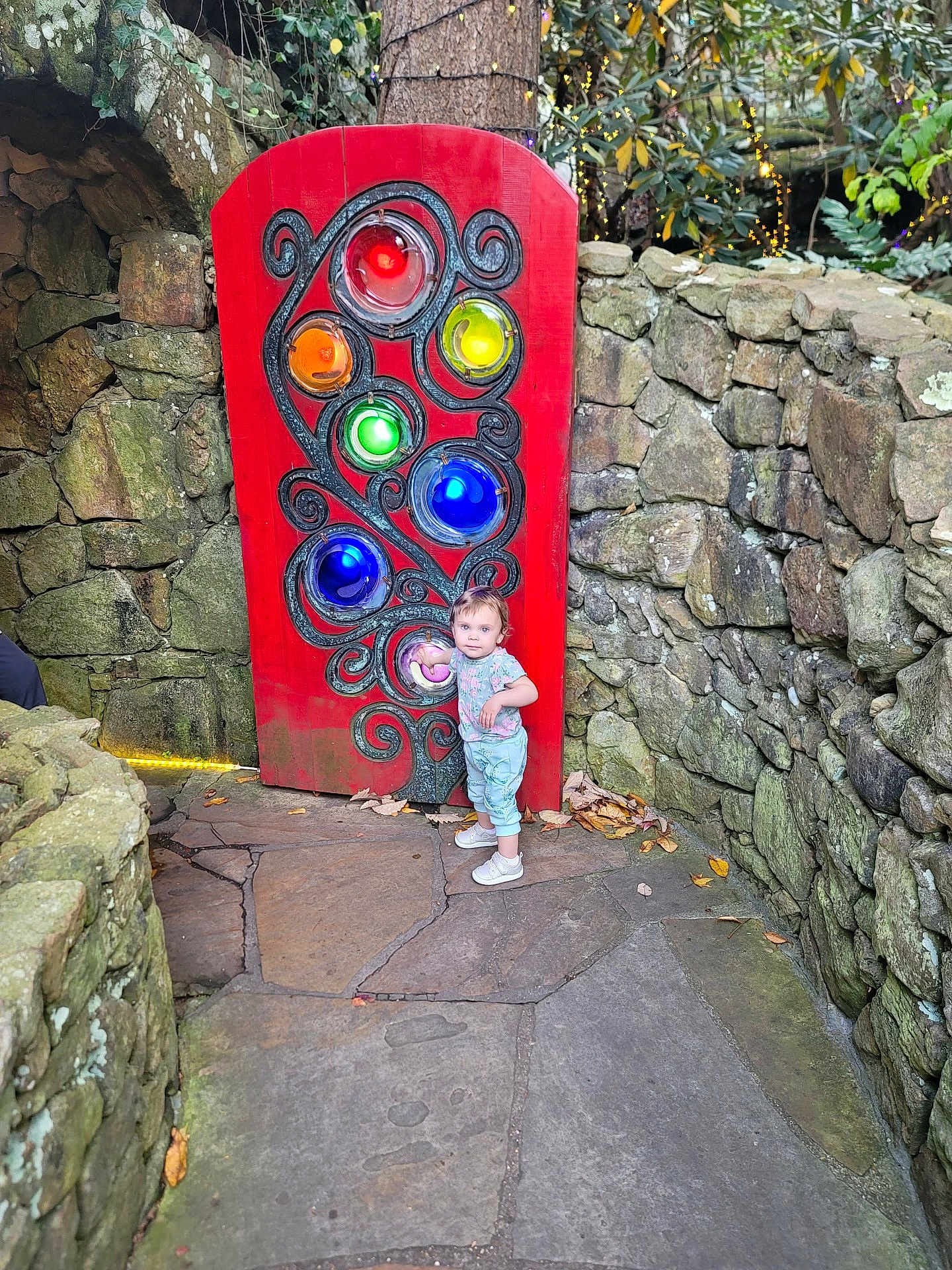Oaklyn joined the competition — help win amazing prizes! child, colorful_lights, curious, decorative, glass_orbs, holiday_lights, leaves, moss, outdoor, pathway, playful, portrait, red_panel, rustic, shoes, standing, stone_wall, toddler, tree, wanderer