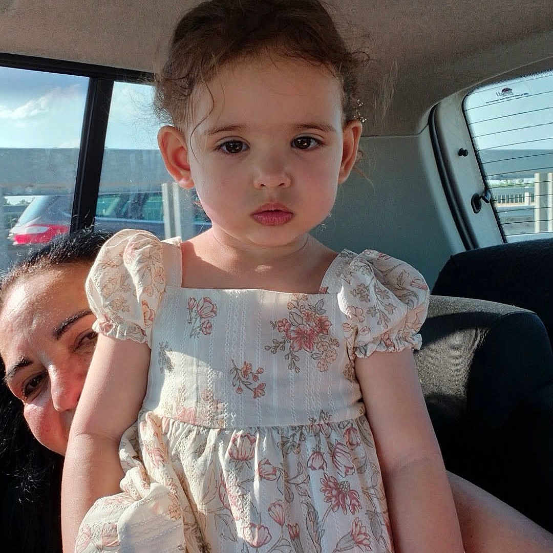 Sofia Maria is registered to the contest to win money with this photo: child, girl, floral_dress, car_interior, woman, peeking, sunlight, serious_expression, portrait, casual, candid, window, seat, road, hair, face, person, clothing, adult, vehicle