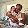 Karson is registered to the contest to win money with this photo: baby, man, smile, tattoo, couch, indoor, family, happy, child, holding, casual_clothing, living_room, person, socks, beard, shorts, tank_top, clock, door, fan