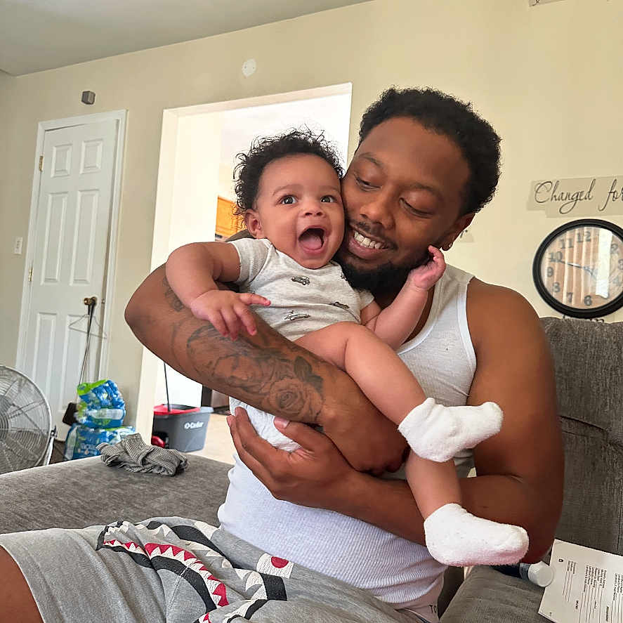 Karson is registered to the contest to win money with this photo: baby, beard, casual_clothing, child, clock, couch, door, family, fan, happy, holding, indoor, living_room, man, person, shorts, smile, socks, tank_top, tattoo