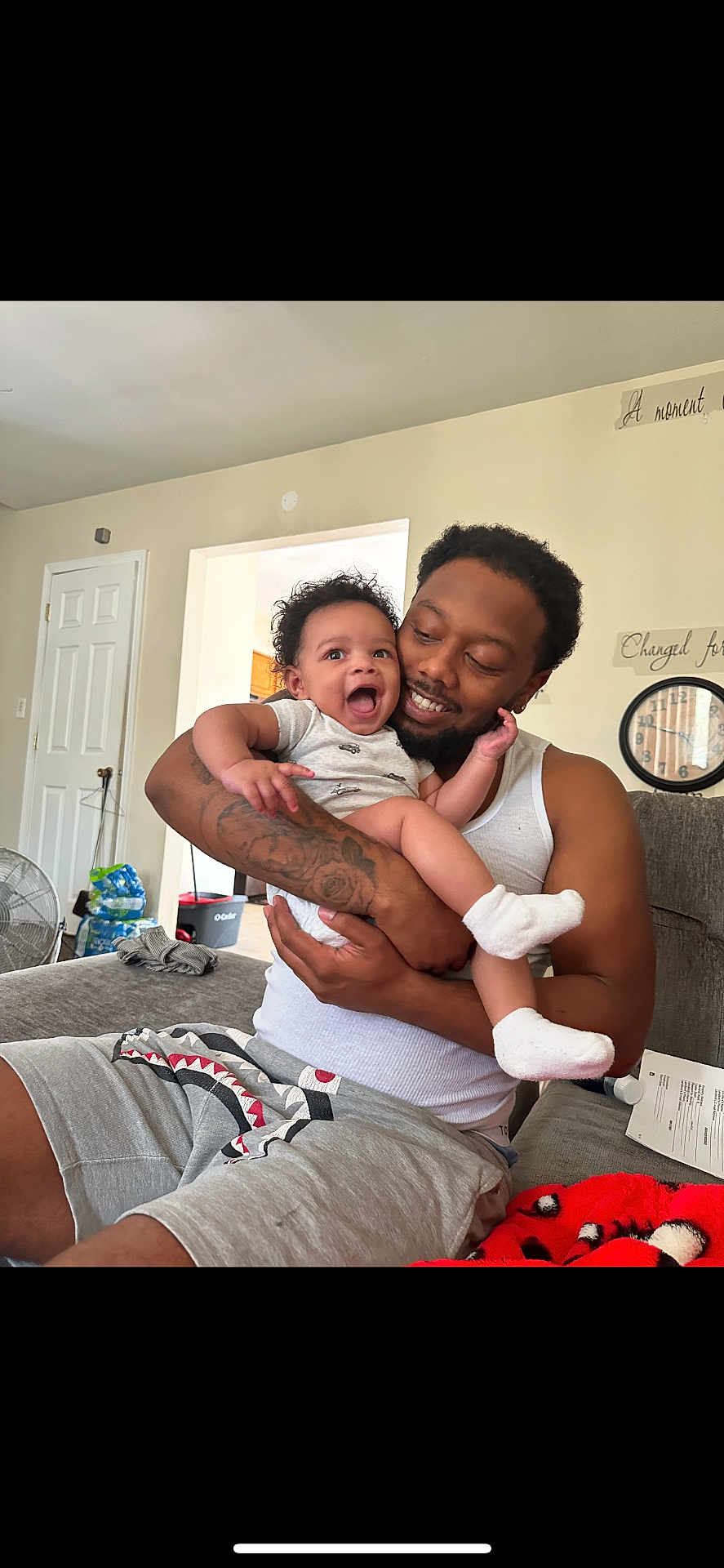 Karson is registered to the contest to win money with this photo: baby, man, smile, tattoo, couch, indoor, family, happy, child, holding, casual_clothing, living_room, person, socks, beard, shorts, tank_top, clock, door, fan