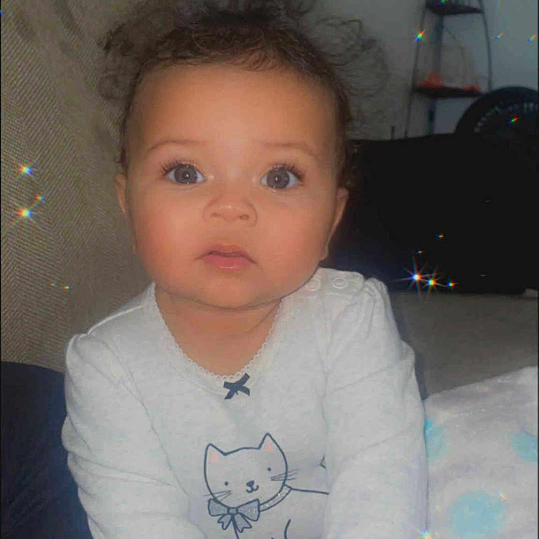 Za'Veha is registered to the contest to win money with this photo: baby, blanket, cat_drawing, child, clothing, couch, cozy, curly_hair, cute, floor, hands, indoor, infant, looking_at_camera, person, portrait, sitting, soft_light, sparkles, white_shirt