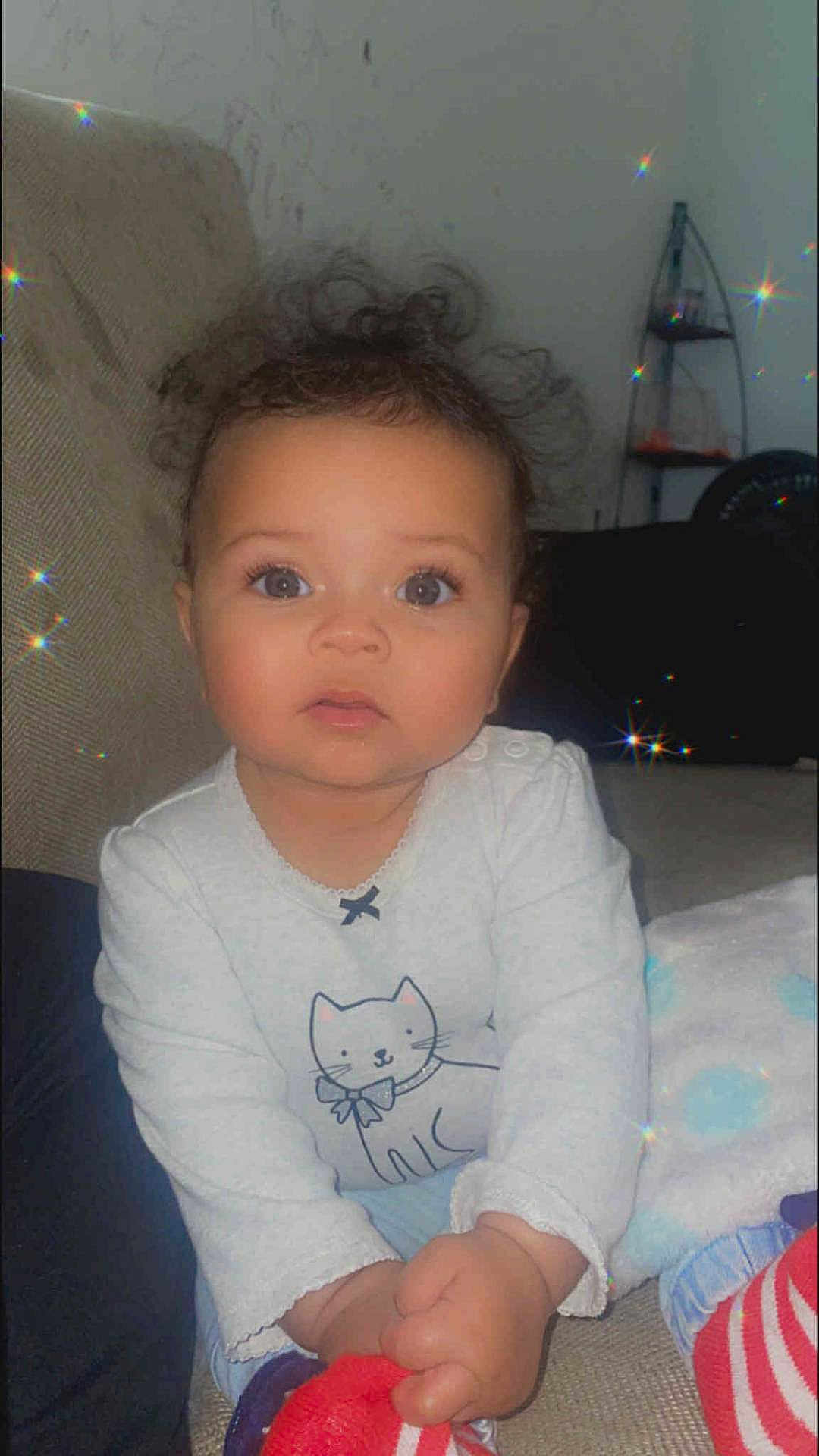 Za'Veha is registered to the contest to win money with this photo: baby, child, curly_hair, indoor, couch, white_shirt, cat_drawing, sitting, cute, sparkles, soft_light, hands, looking_at_camera, infant, cozy, person, portrait, clothing, floor, blanket